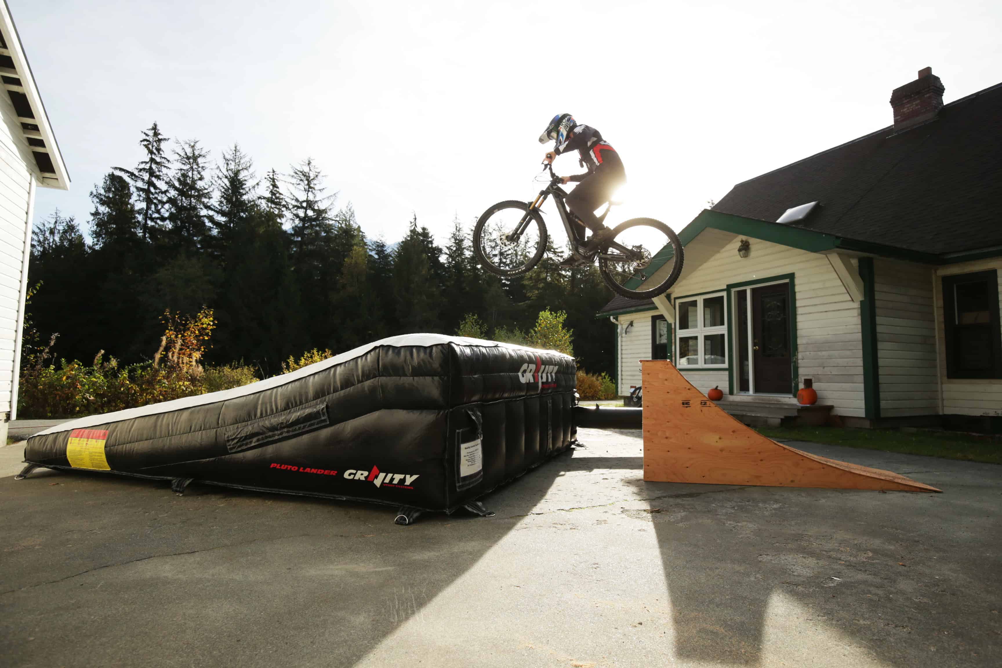The Pluto Lander airbag seen from the side as a rider launches from a Gravity Stunts ramp, framed by trees and a house in the background.