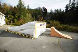 Mountain biker launching off a Gravity Stunts ramp onto the Lunar Lander airbag, with visible dimensions, trees, and a mountain in the background.