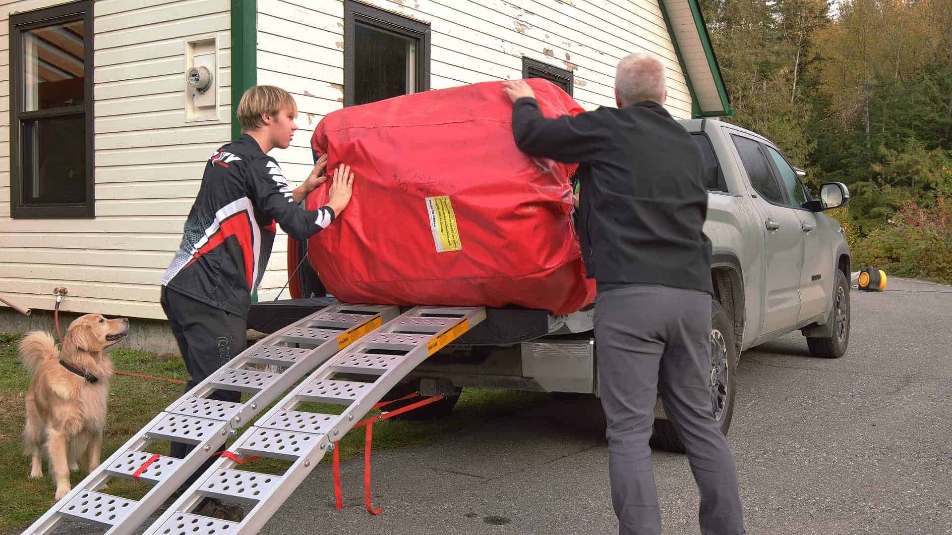 Two mountainbikers and a very helpful dog snoopervising loading the landing ramp or mat on a metal ramp into a pickup.